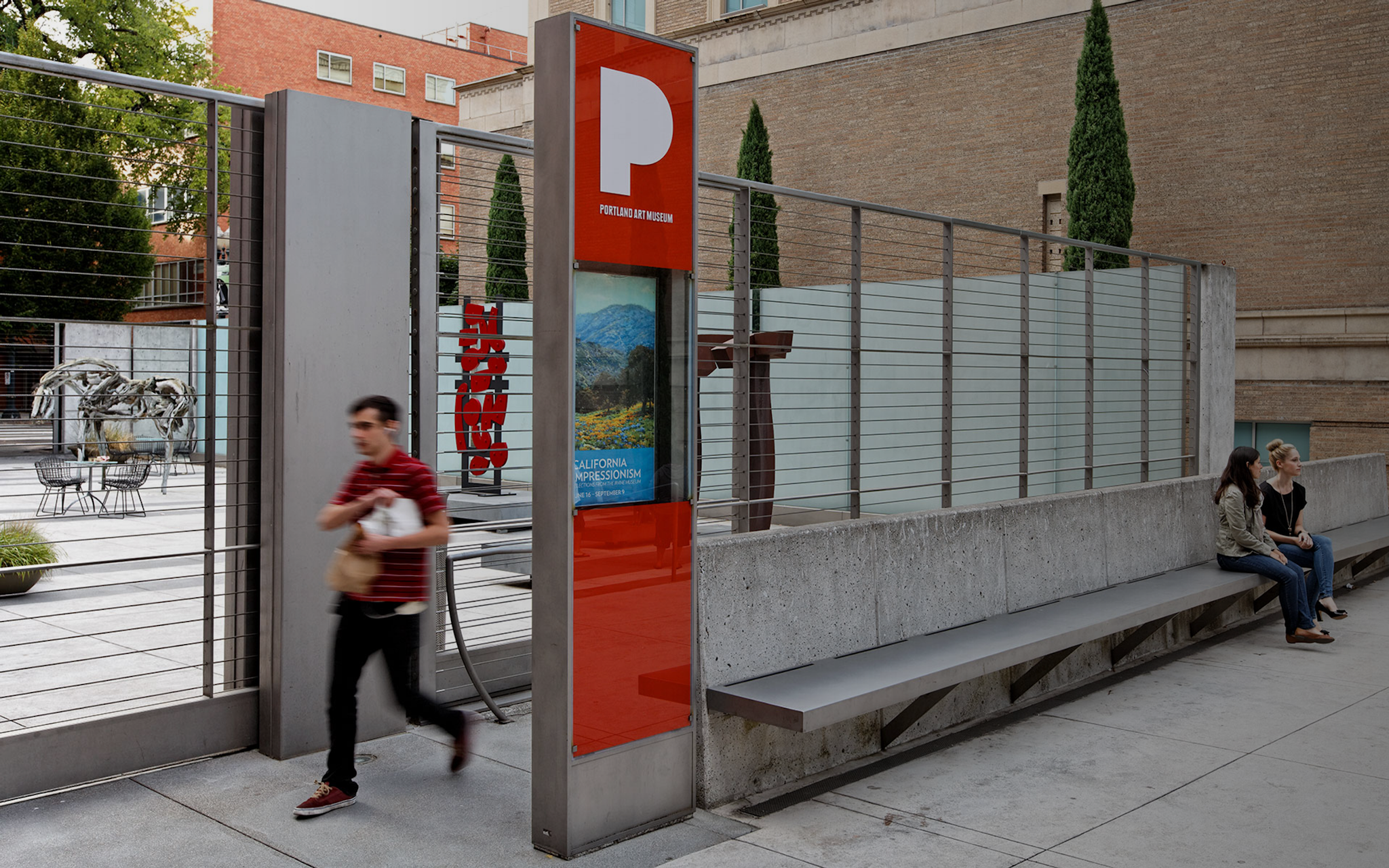 Person walking next to a Portland Art Museum kiosk that is beside a long concrete bench in front of a sculpture plaza