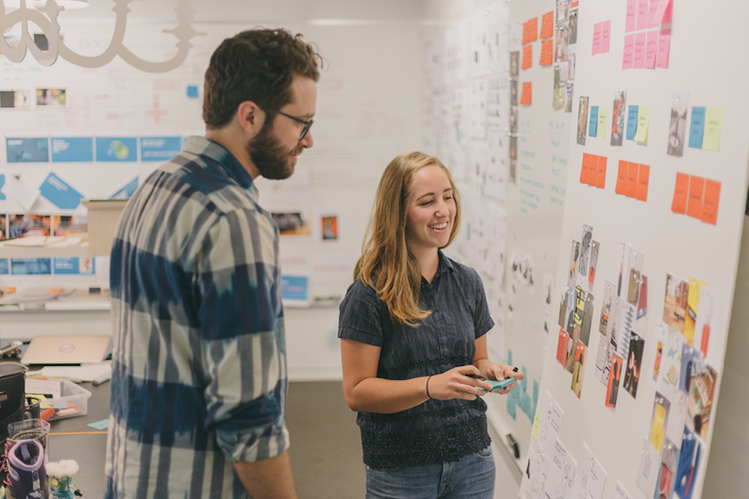 Two people smiling and looking at a whiteboard with sticky notes on it