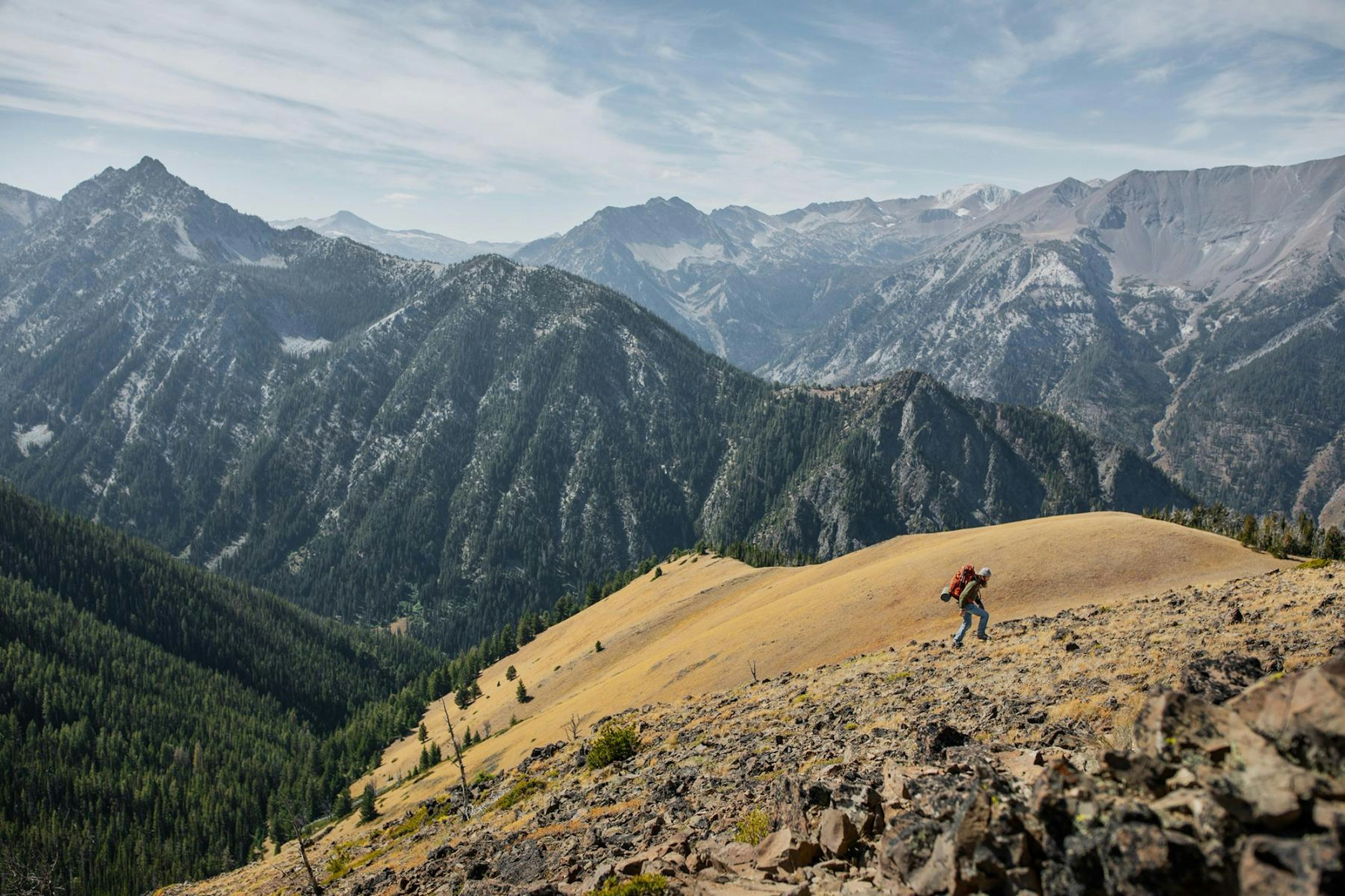 Person wearing a large backpack hiking up a mountain side in front of a mountain range covered in trees