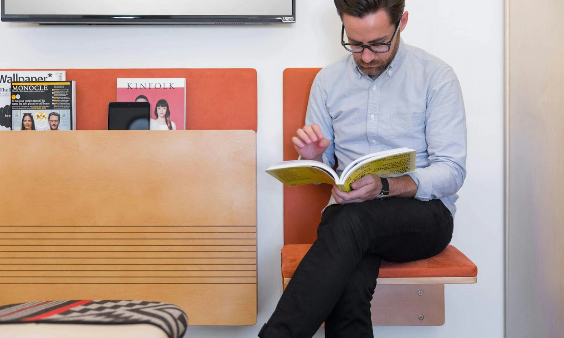 Man sitting on a JumpSeat in a modern waiting area reading a magazine, with magazines displayed on the wall behind him.