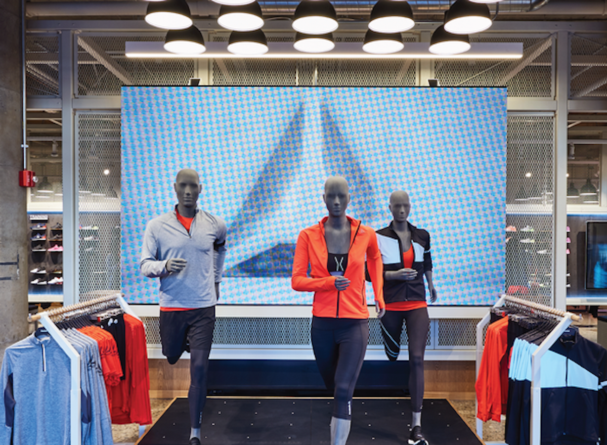 Three mannequins in running positions wearing sports attire in front of a large digital display