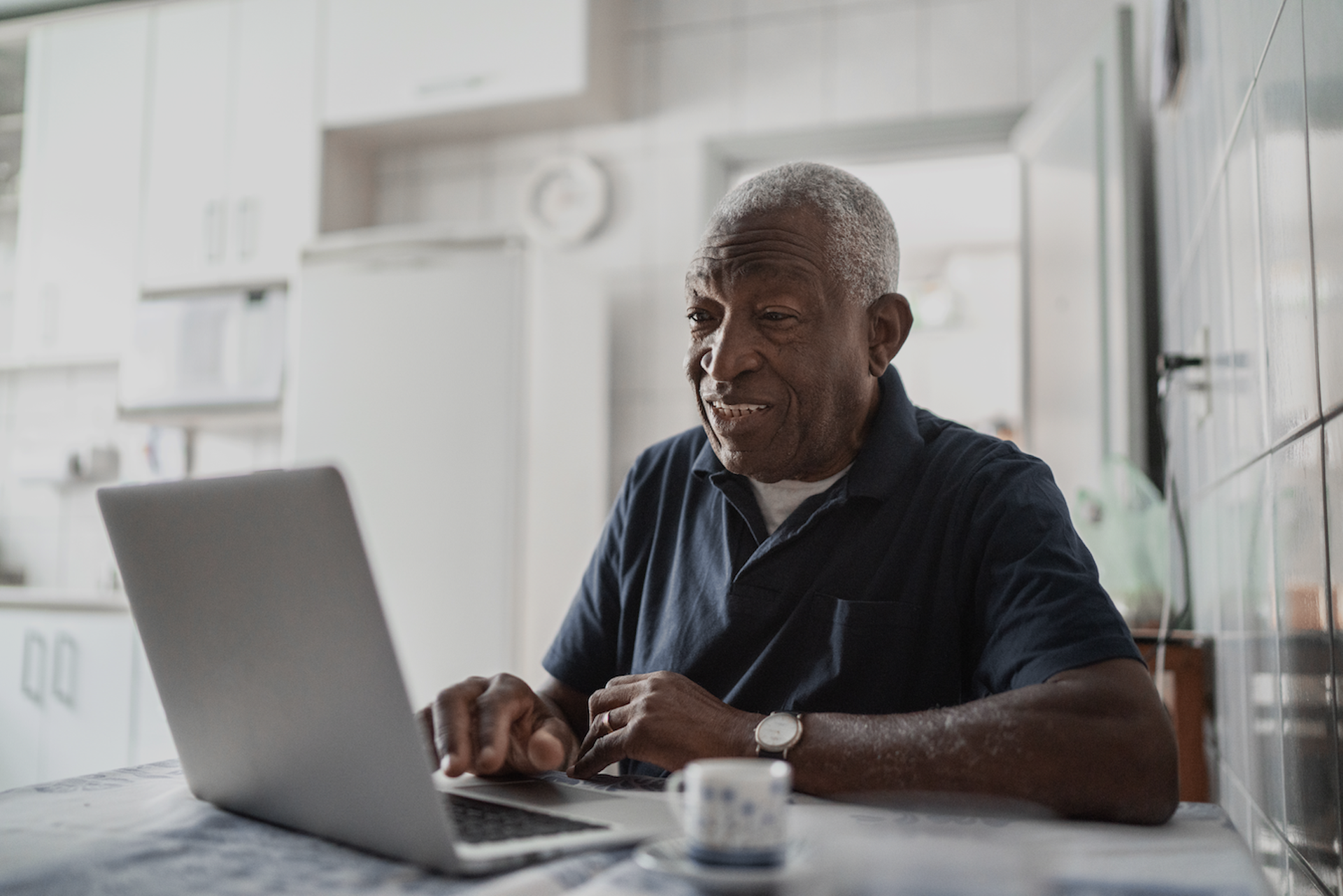 Elderly man sitting at a kitchen table, smiling and looking at an open laptop