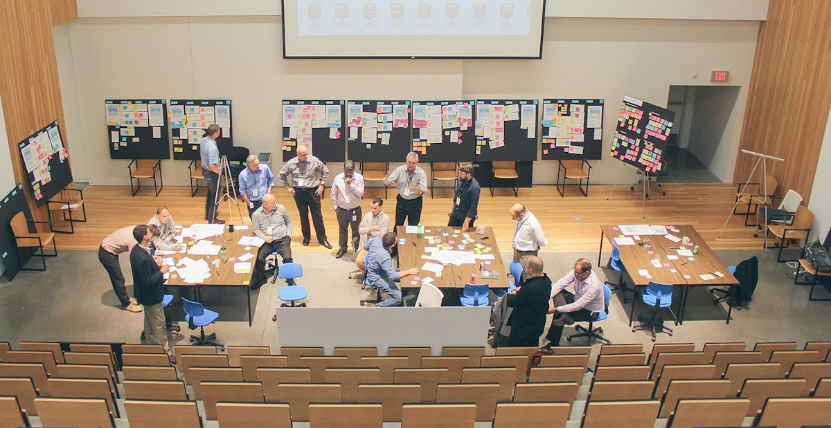 Several people having conversations during a workshop, in front of five foam blackboards that have sticky notes and sheets of paper attached to them